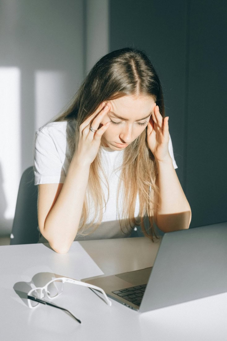 A young woman with a headache holding her head, sitting in front of a laptop.